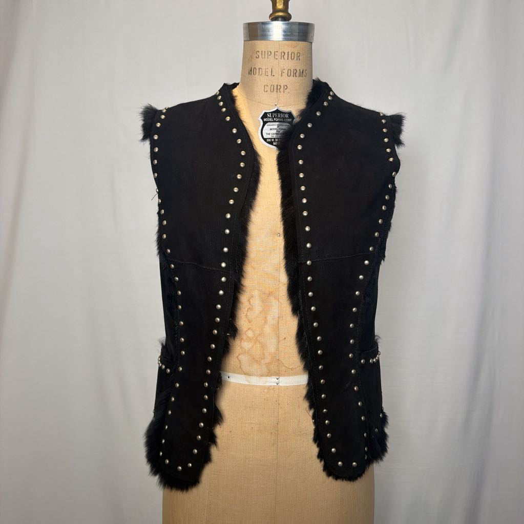 A black suede vest with silver button embellishments and black fur on the inside fluffing out. It's worn on an antique mannequin that sits in front of a white fabric wall.