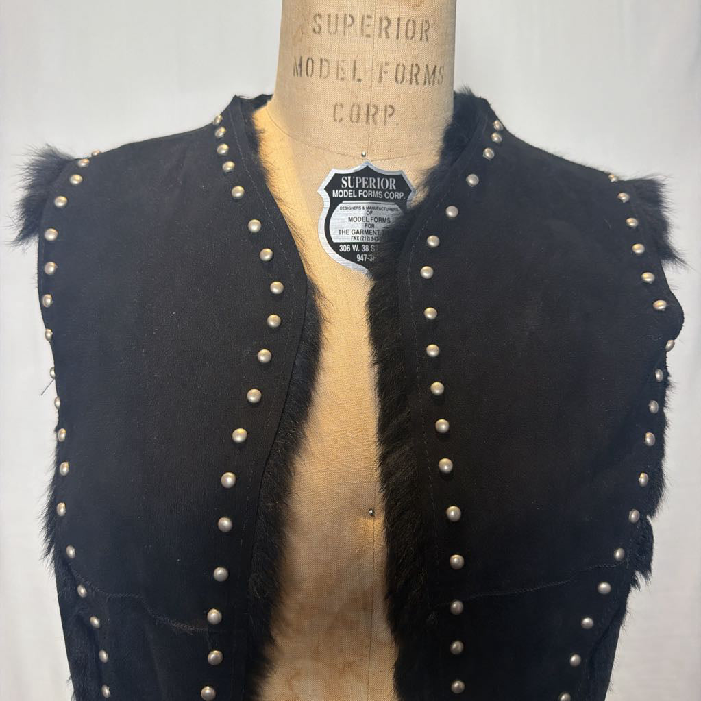 A black suede vest with silver button embellishments and black fur on the inside fluffing out. It's worn on an antique mannequin that sits in front of a white fabric wall.