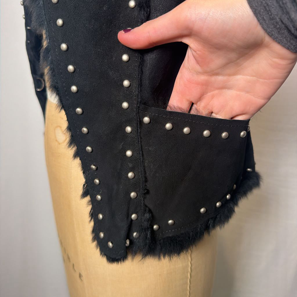 A black suede vest with silver button embellishments and black fur on the inside fluffing out. It's worn on an antique mannequin that sits in front of a white fabric wall.