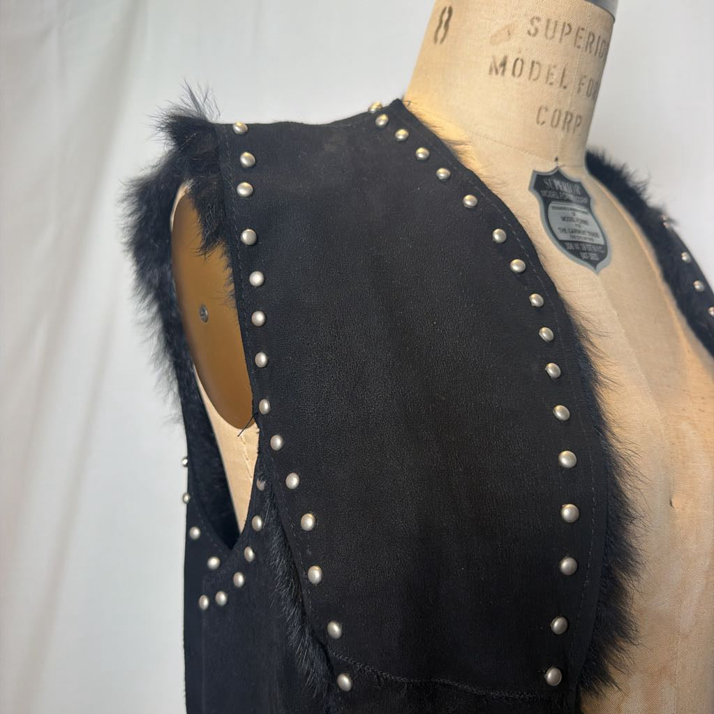 A black suede vest with silver button embellishments and black fur on the inside fluffing out. It's worn on an antique mannequin that sits in front of a white fabric wall.