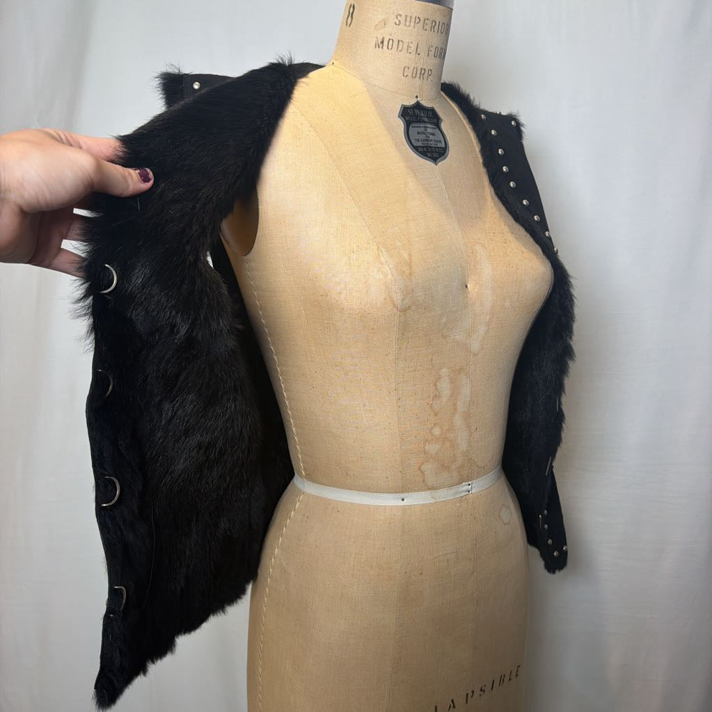 A black suede vest with silver button embellishments and black fur on the inside fluffing out. It's worn on an antique mannequin that sits in front of a white fabric wall.