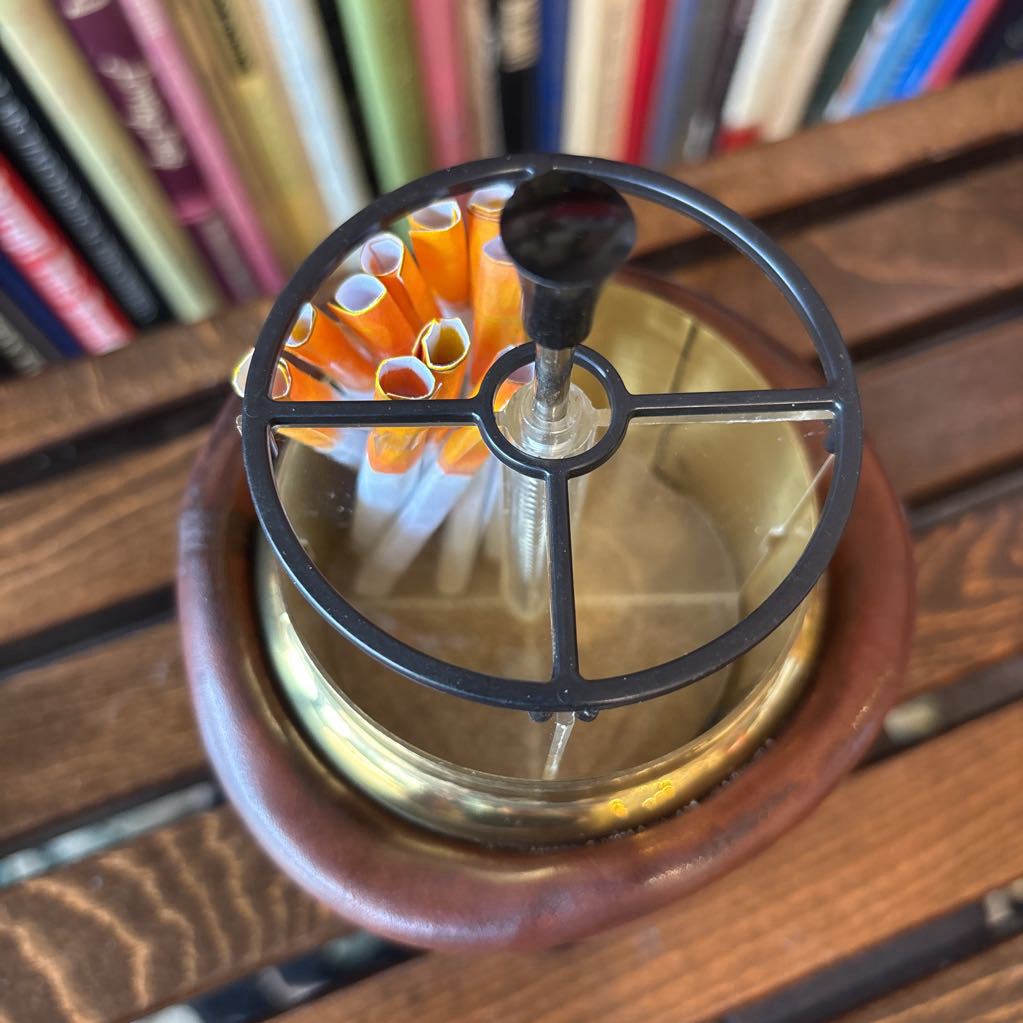 A brass and black metal cigarette holder with orange and white filter tips, set inside a brown leather-like holder on a wooden table with a blurred bookshelf background.