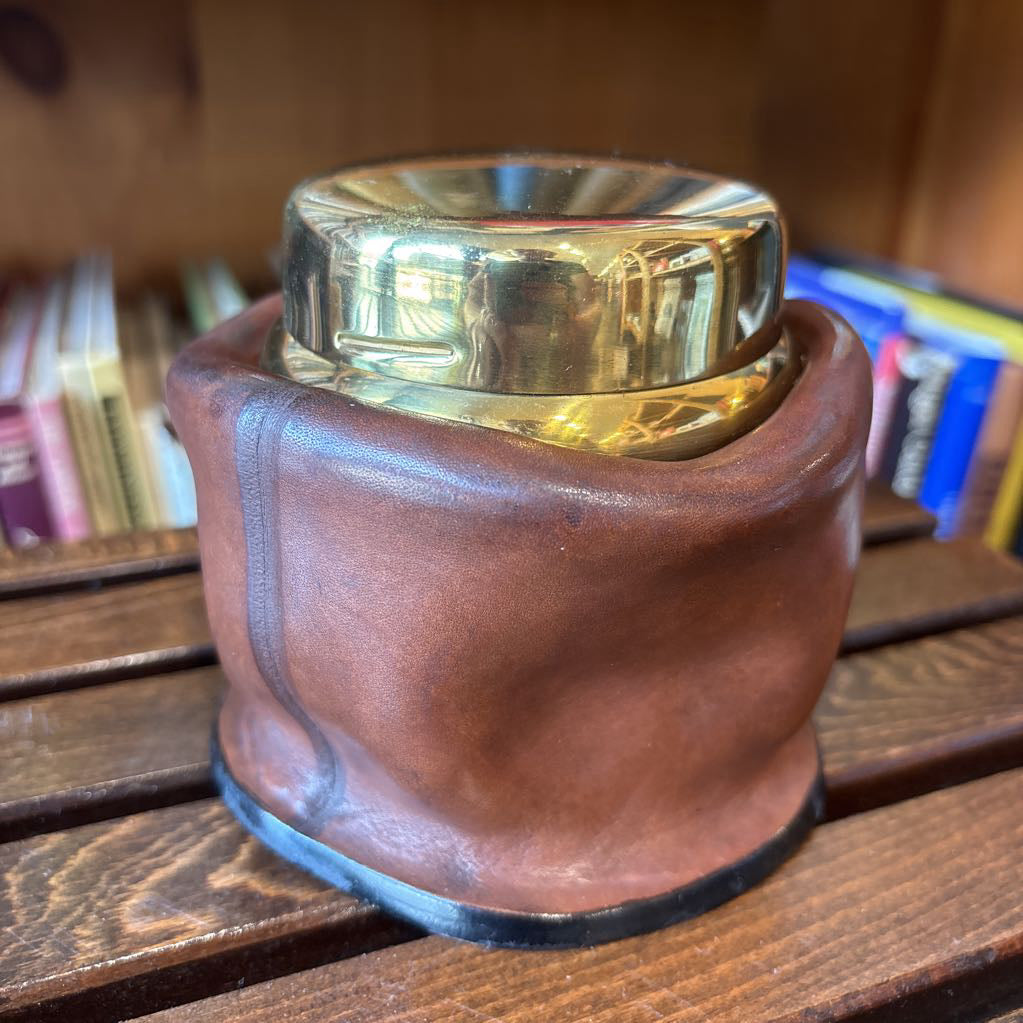 A brass and leather round object on a wooden slat bench in front of books.