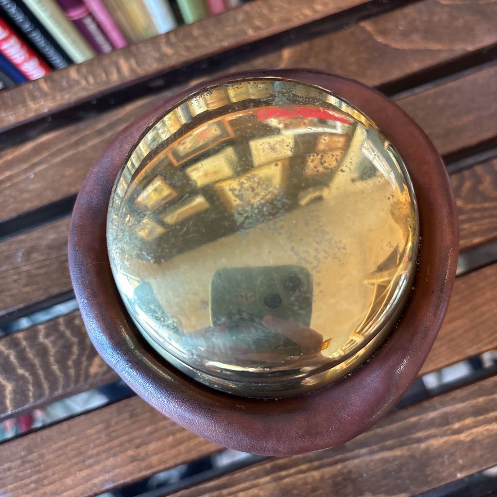 A brass and leather round object on a wooden slat bench in front of books.