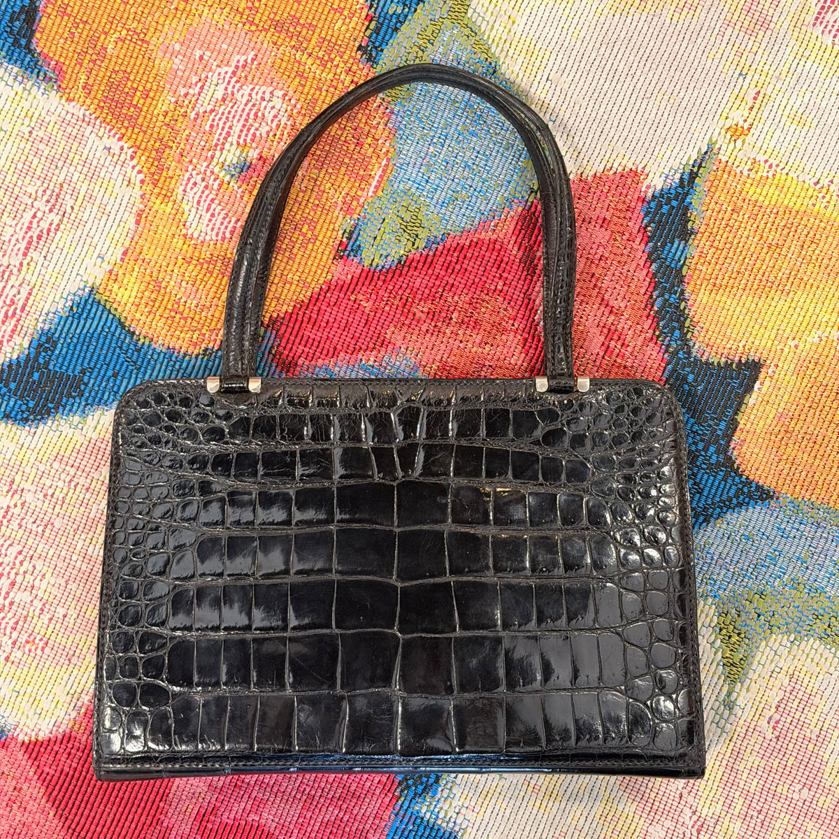 A black crocodile-embossed leather handbag with a structured rectangular shape and double top handles, displayed on a colorful floral woven textile background.