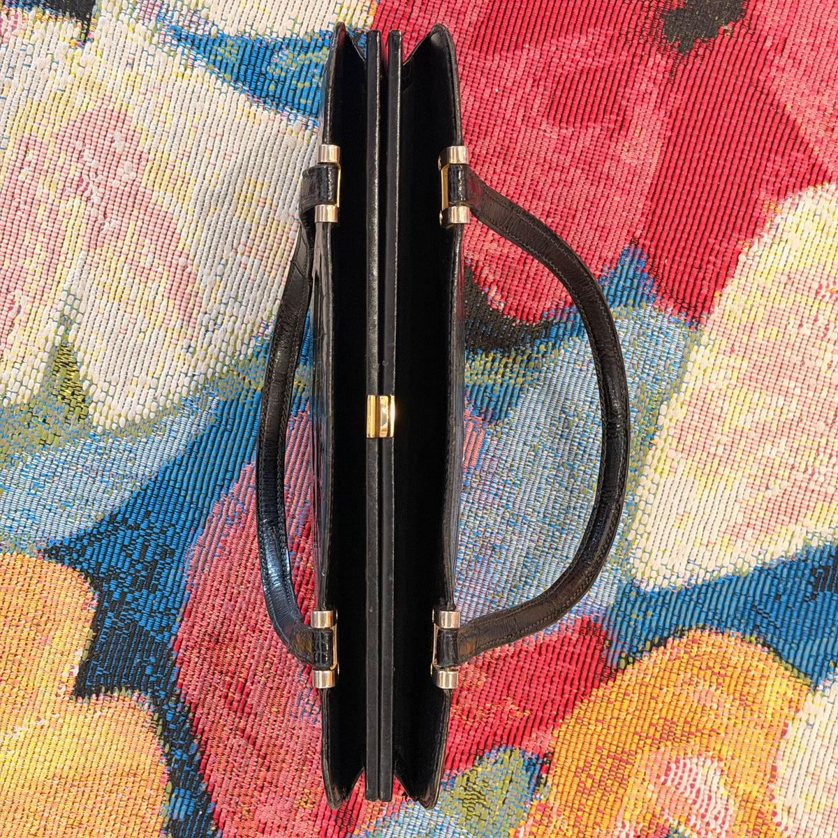 A black leather handbag shown closed from the side, featuring a structured double-gusset design, a top handle, and gold-tone hardware, set against a colorful floral woven textile background.