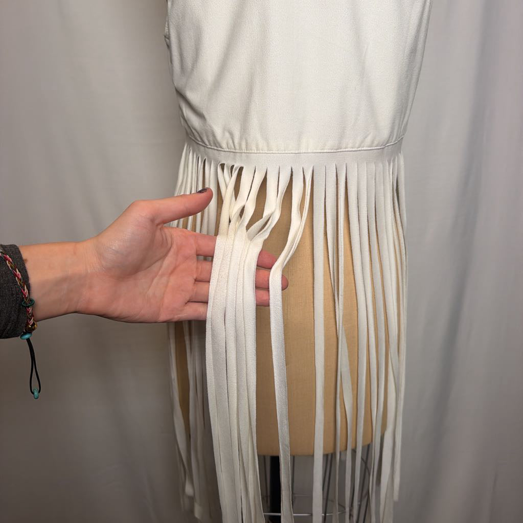A white vest with long fringe on an antique mannequin in front of a white fabric wall.