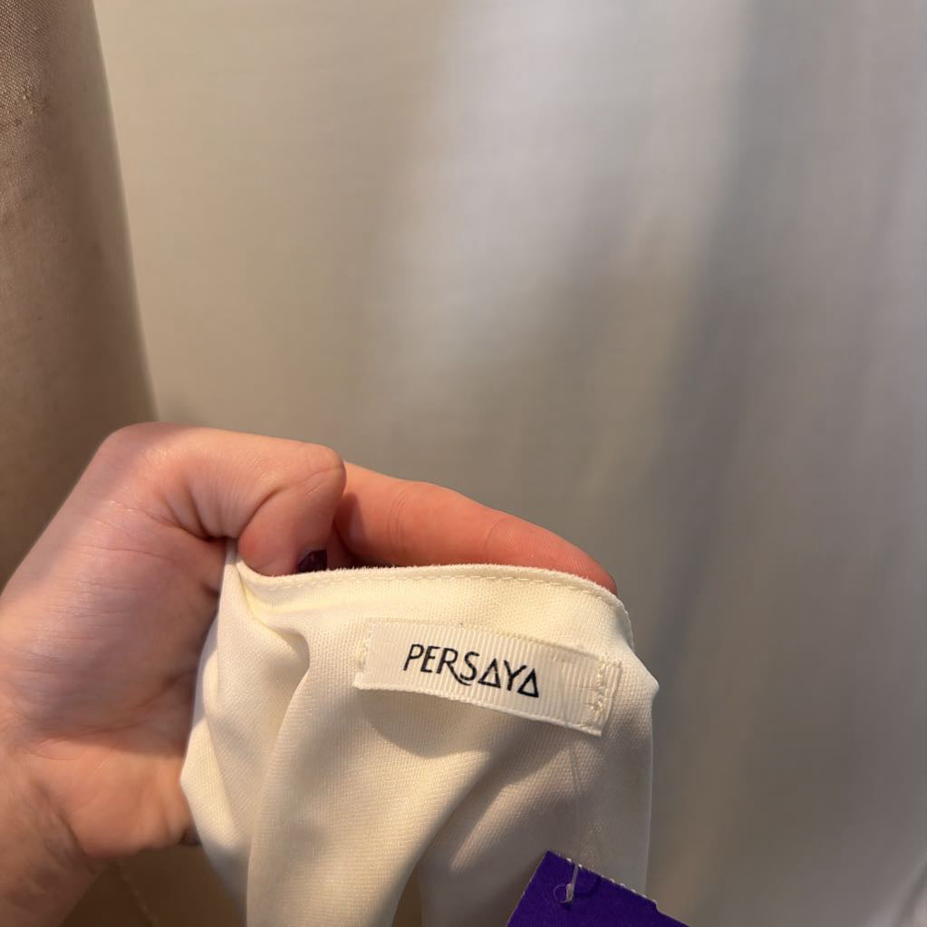 A hand holding an interior brand tag that says "Persaya" on a white article of clothing in front of a white wall.