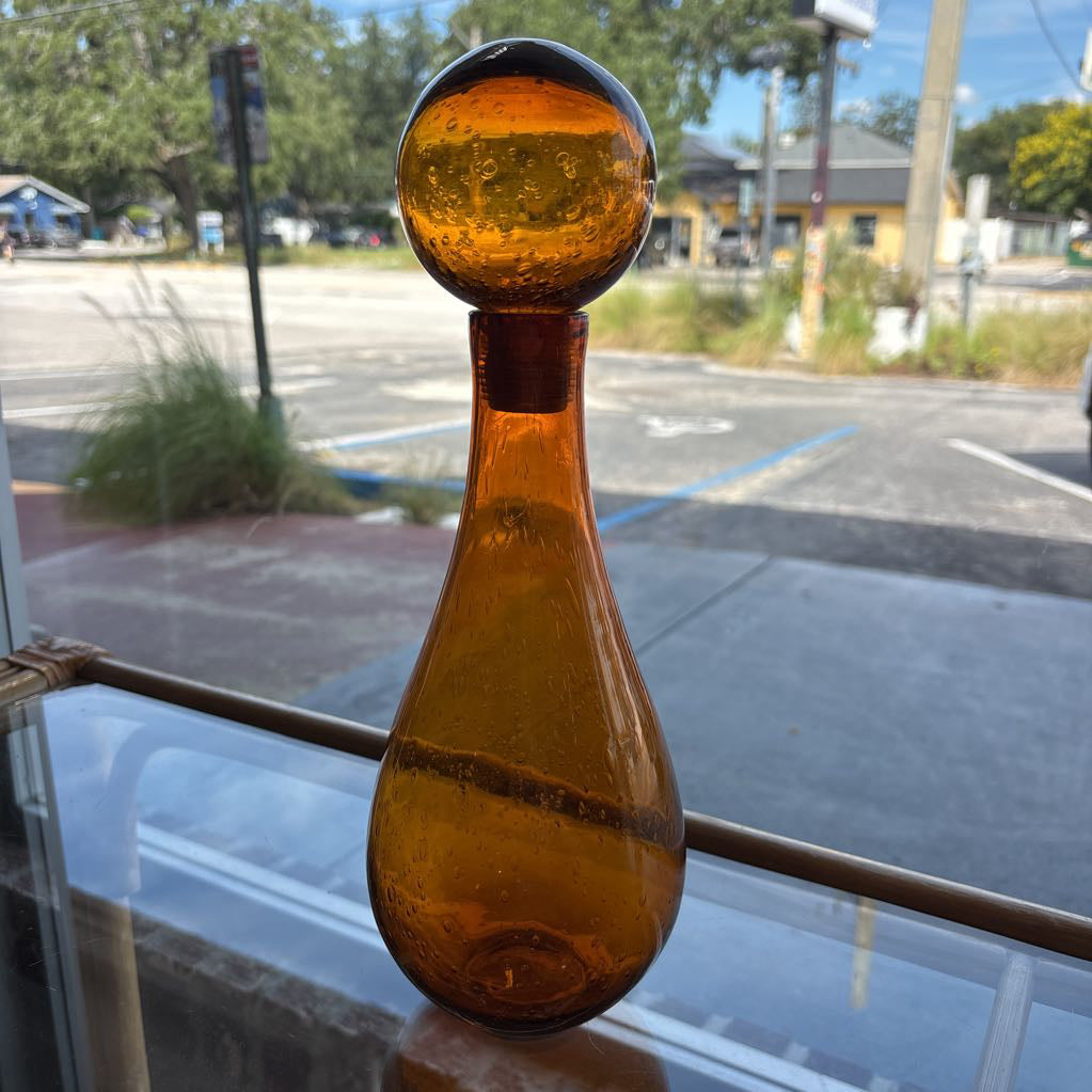 An amber blown glass decanter with a drop shape body and a spherical stopper. Its sitting on a glass table in front of a parking lot.