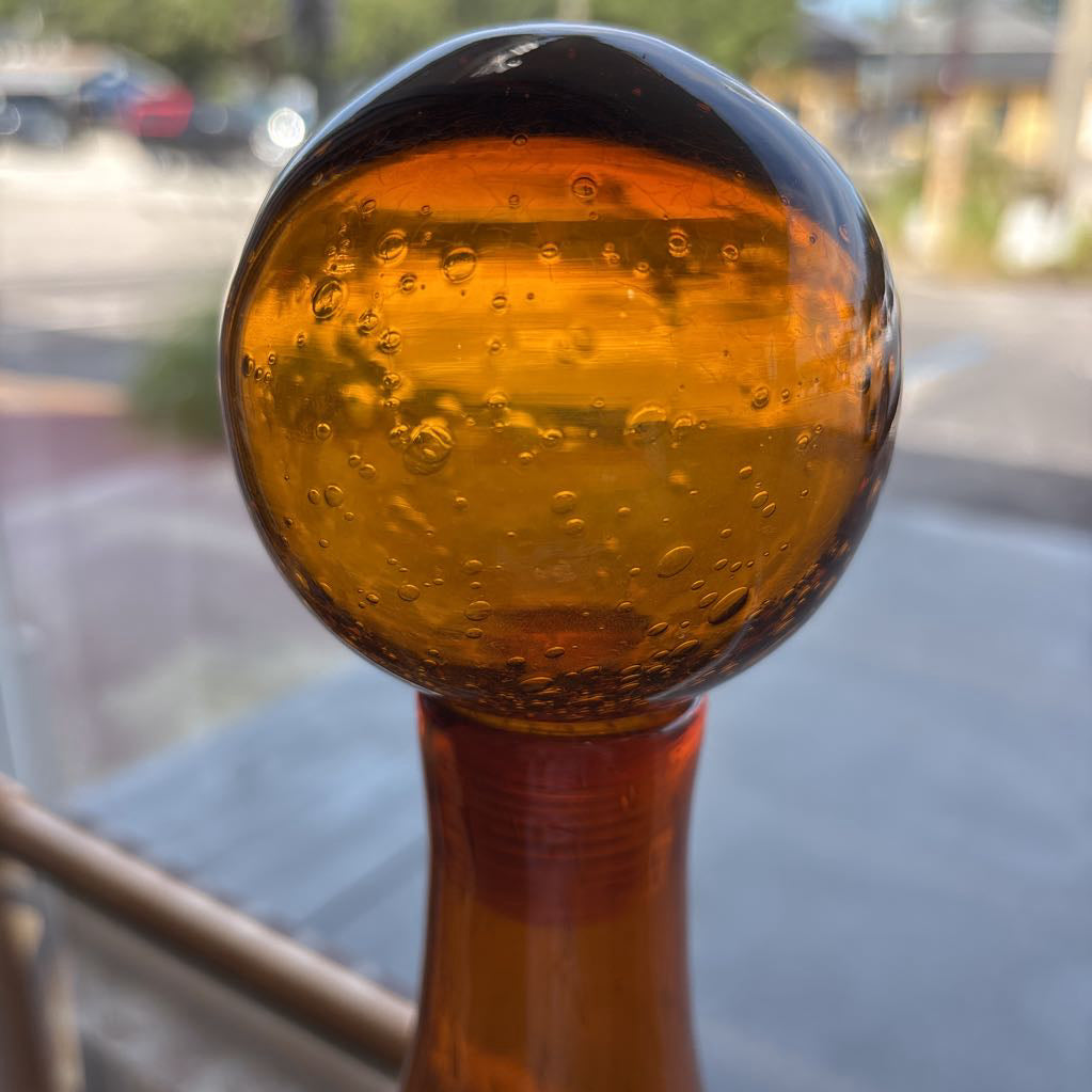 An amber blown glass decanter with a drop shape body and a spherical stopper. Its sitting on a glass table in front of a parking lot.