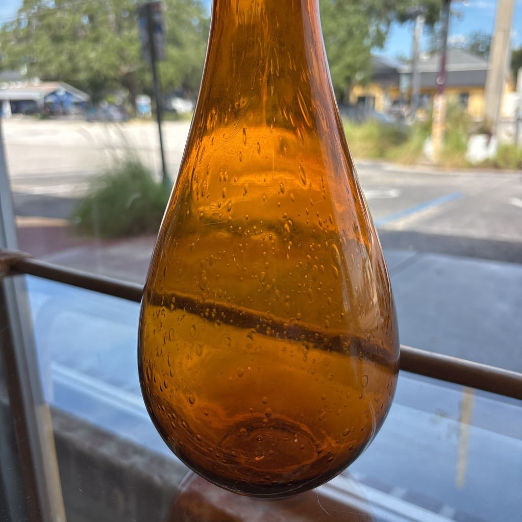 An amber blown glass decanter with a drop shape body and a spherical stopper. Its sitting on a glass table in front of a parking lot.