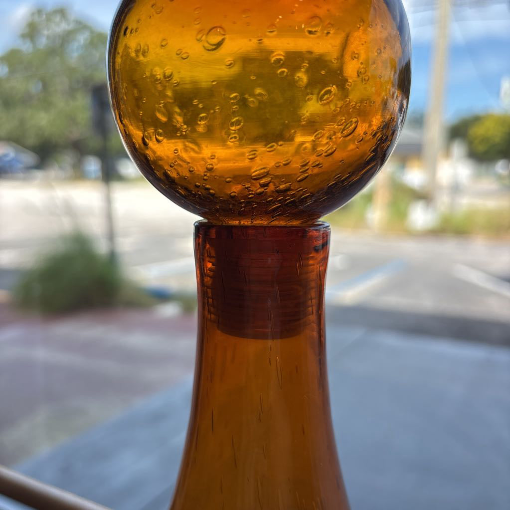 An amber blown glass decanter with a drop shape body and a spherical stopper. Its sitting on a glass table in front of a parking lot.