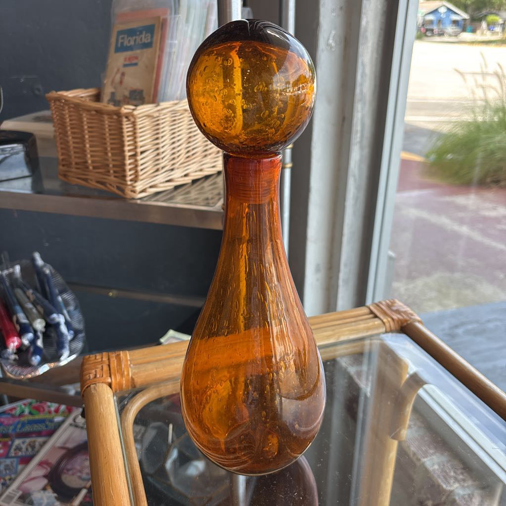 An amber blown glass decanter with a drop shape body and a spherical stopper. Its sitting on a glass table in front of a parking lot.