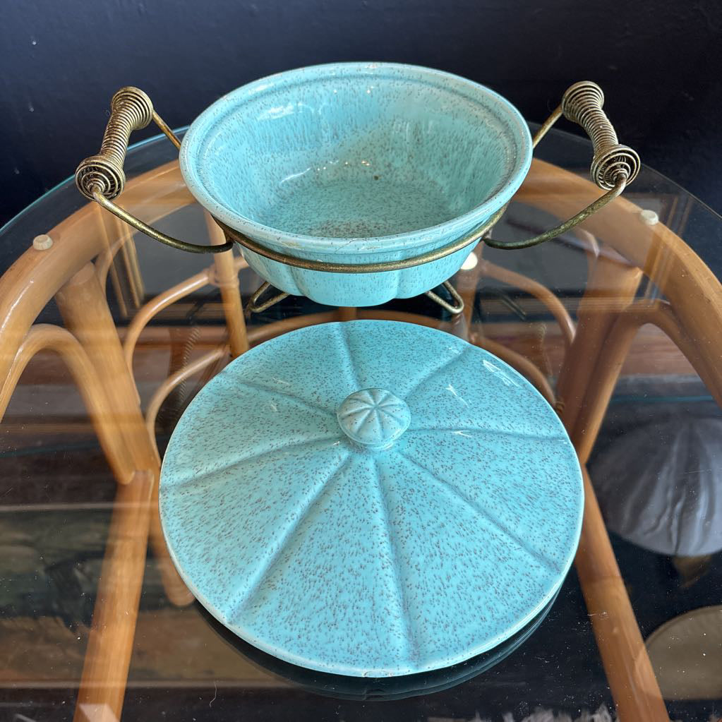 A ceramic turquoise serving bowl and lid in a brass wire caddy with handles on a glass table in front of a black wall.