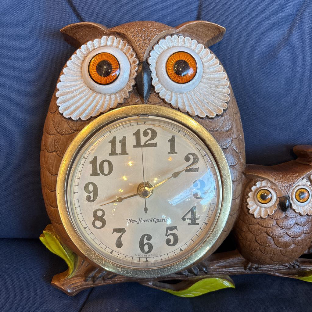 A vintage clock of 3 owls with wide eyes sitting on a branch and the clock face in the largest owl. This clock is on a blue cushion.