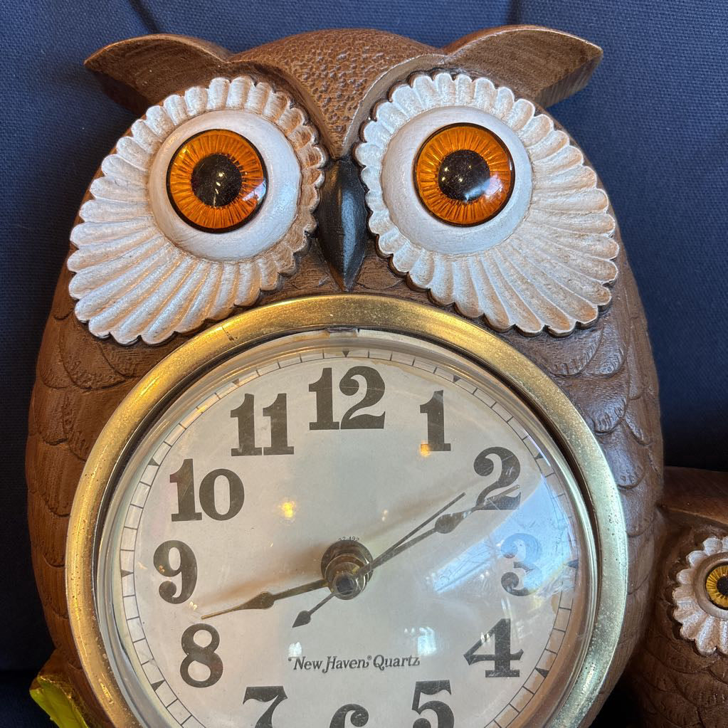 A vintage clock of 3 owls with wide eyes sitting on a branch and the clock face in the largest owl. This clock is on a blue cushion.
