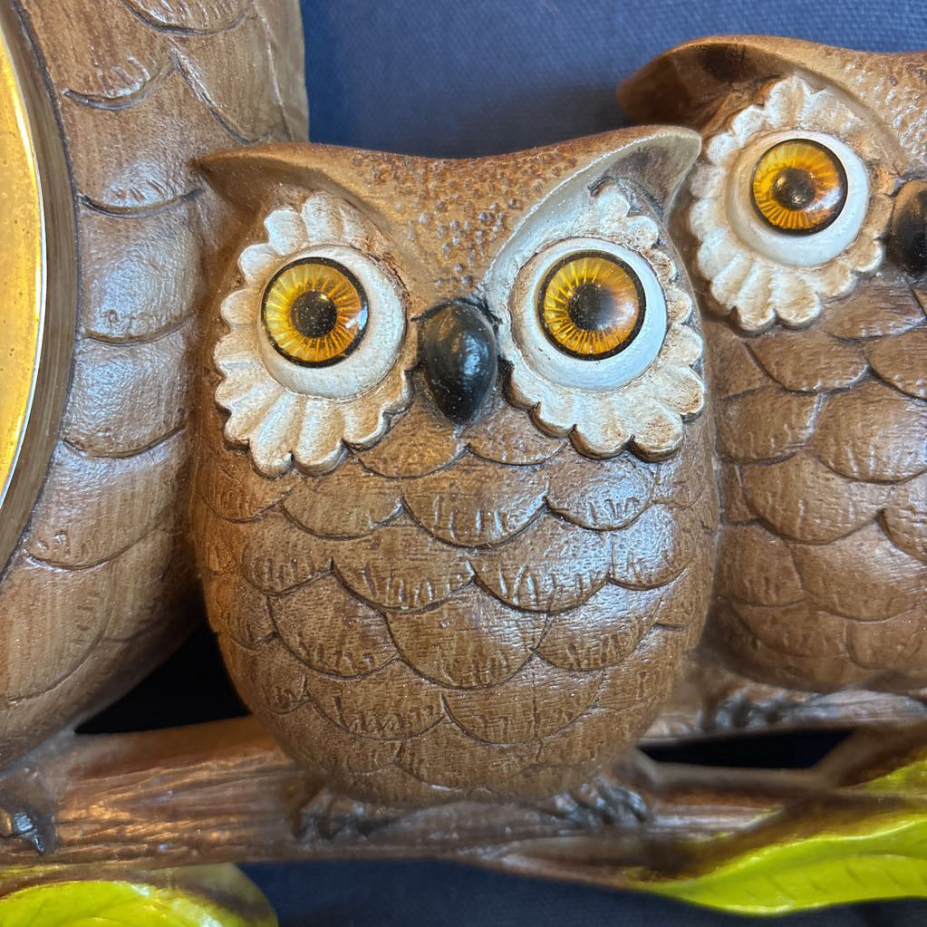 A vintage clock of 3 owls with wide eyes sitting on a branch and the clock face in the largest owl. This clock is on a blue cushion.