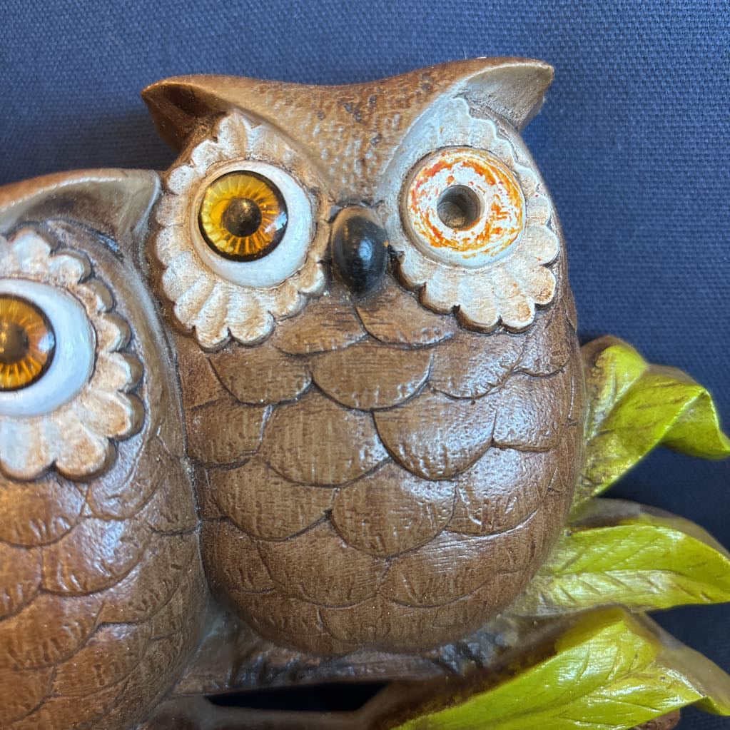 A vintage clock of 3 owls with wide eyes sitting on a branch and the clock face in the largest owl. This clock is on a blue cushion.