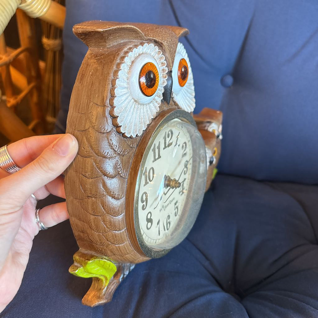 A vintage clock of 3 owls with wide eyes sitting on a branch and the clock face in the largest owl. This clock is on a blue cushion.