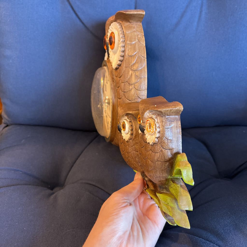 A vintage clock of 3 owls with wide eyes sitting on a branch and the clock face in the largest owl. This clock is on a blue cushion.