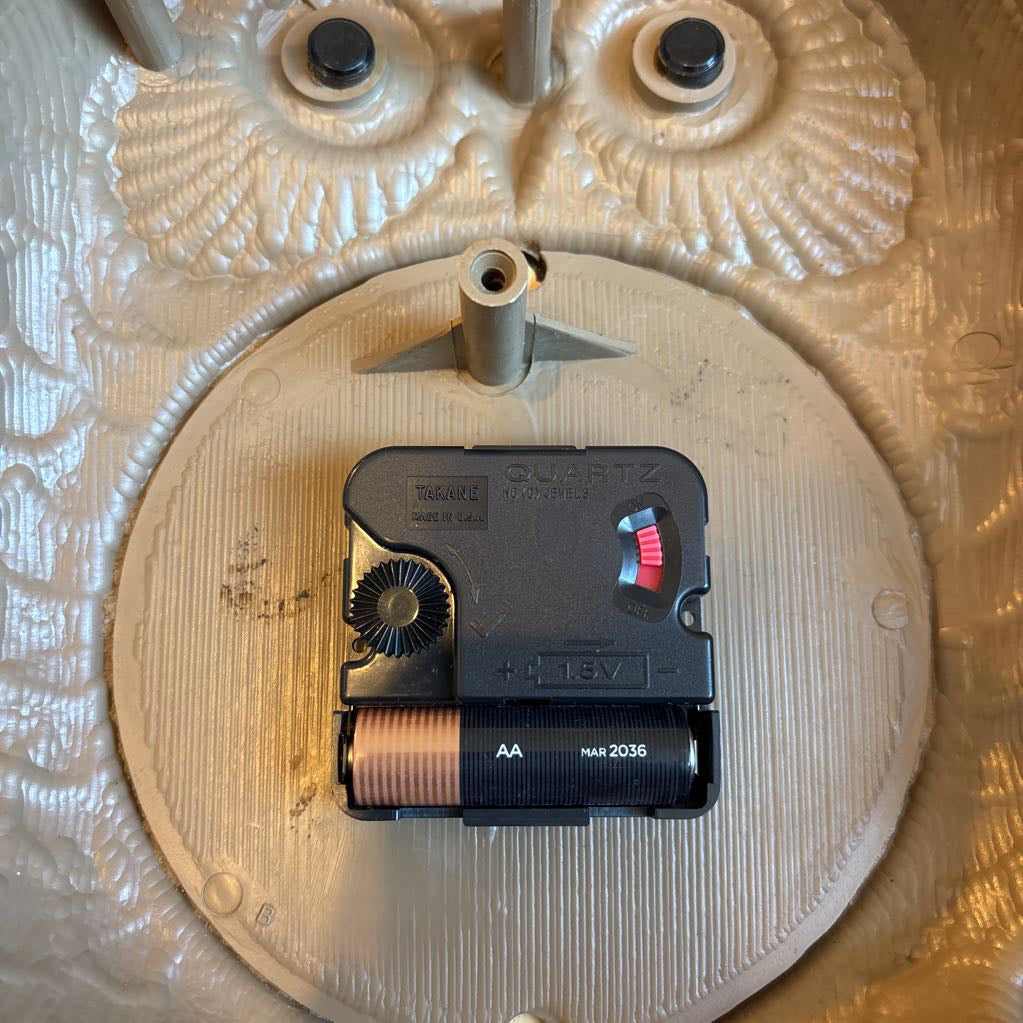 A vintage clock of 3 owls with wide eyes sitting on a branch and the clock face in the largest owl. This clock is on a blue cushion.