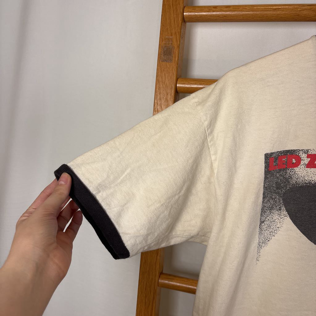 A white vintage style short sleeve t-shirt with an image of a Led Zeppelin album cover and the band's name in red letters. The shirt hangs on a wooden rack against a white background.