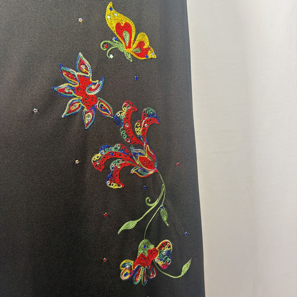 A black maxi dress with a butterfly and floral embroidery at the bottom. This dress is on a mannequin in front of a white wall.