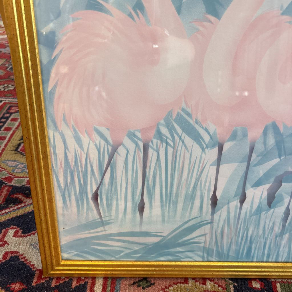 A print of three pink flamingos in a tropical setting in a gold frame in a room setting.