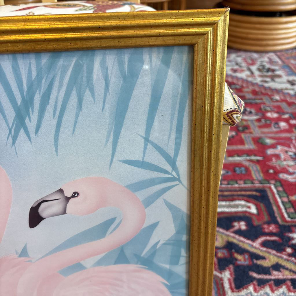 A print of three pink flamingos in a tropical setting in a gold frame in a room setting.
