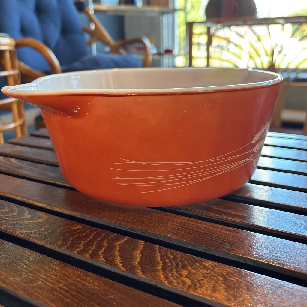 An orange pyrex casserole dish with the imagery of stalks of wheat across the exterior. The dish sits on a wooden slat bench in a room setting.