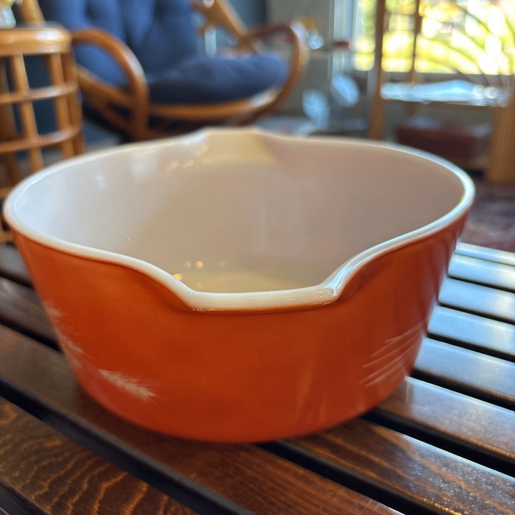 An orange pyrex casserole dish with the imagery of stalks of wheat across the exterior. The dish sits on a wooden slat bench in a room setting.