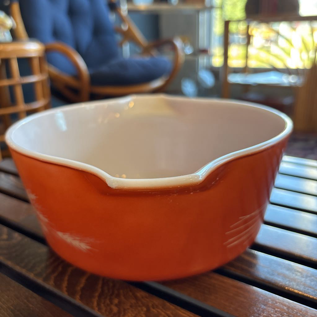 An orange pyrex casserole dish with the imagery of stalks of wheat across the exterior. The dish sits on a wooden slat bench in a room setting.