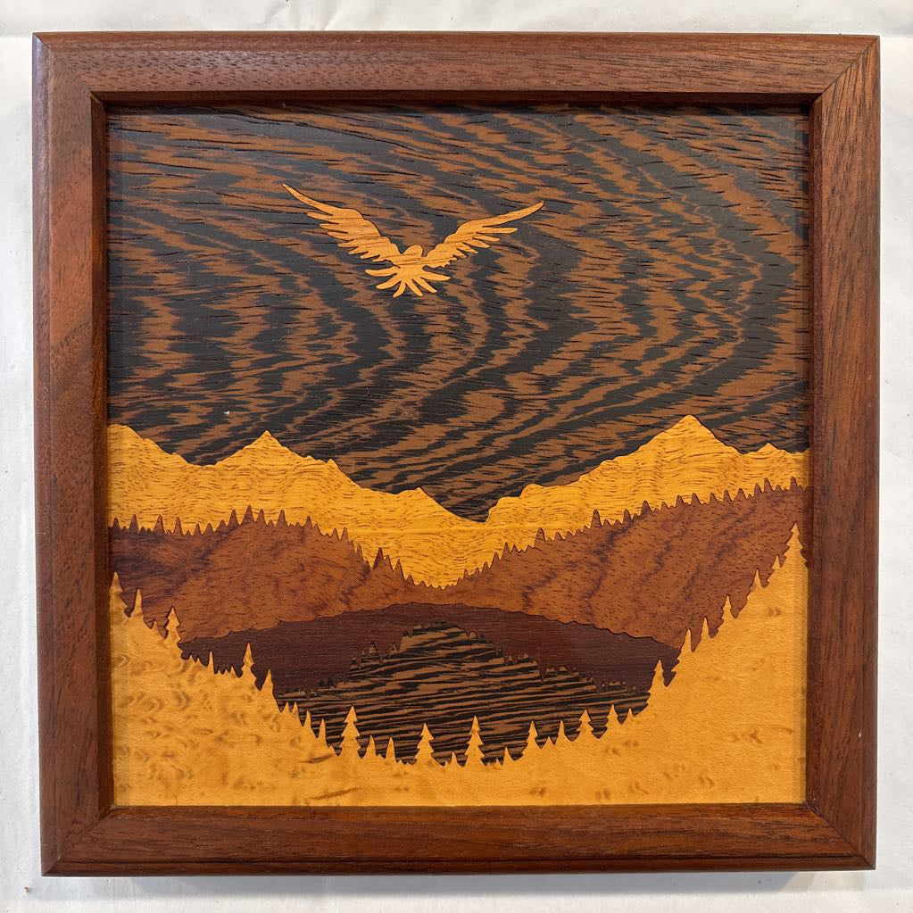 A square framed wooden inlaid artwork of a bird flying over mountains and forests with different colors and textures of wood.
