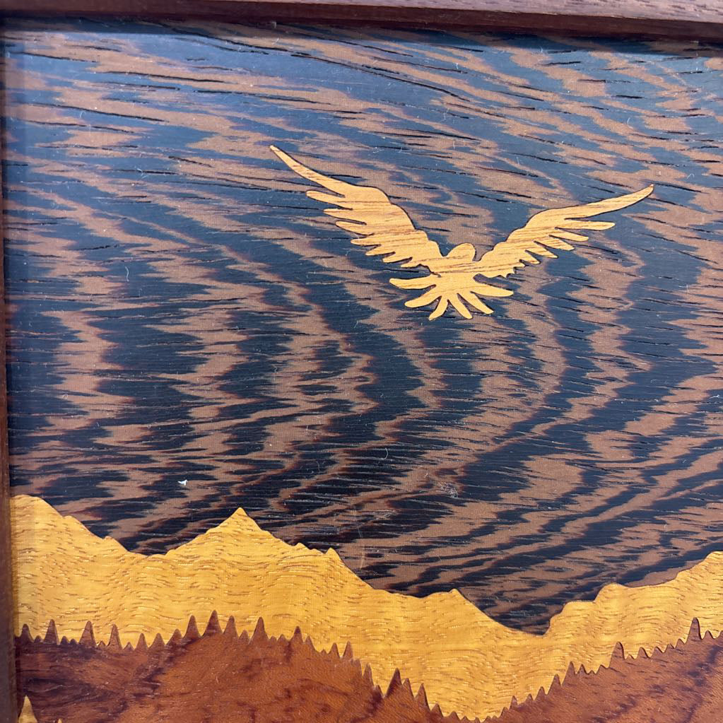 A square framed wooden inlaid artwork of a bird flying over mountains and forests with different colors and textures of wood.