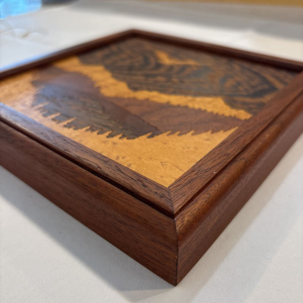A square framed wooden inlaid artwork of a bird flying over mountains and forests with different colors and textures of wood.