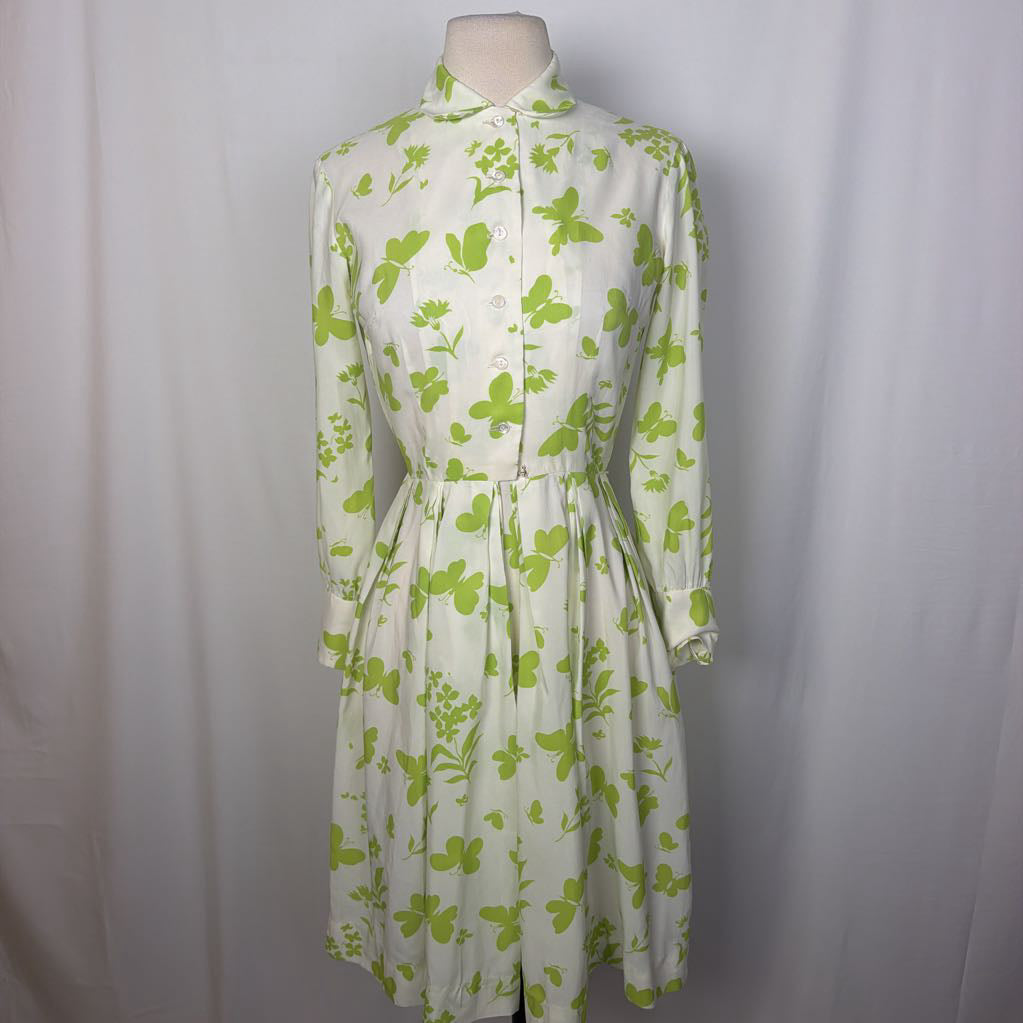 A long-sleeve collared dress displayed on a mannequin torso against a light gray fabric backdrop. The dress has an off-white base with a bright green floral and leaf print, a button front, and a self-tie waist belt.