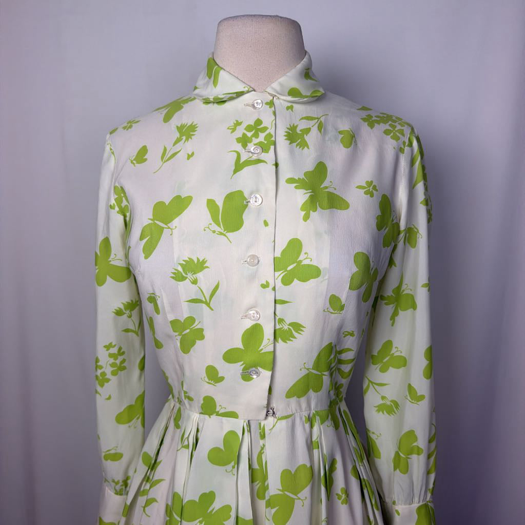 A vintage-style long-sleeve dress displayed on a mannequin torso, featuring a light cream base with a green clover and floral print, a pointed collar, and a button-front bodice.