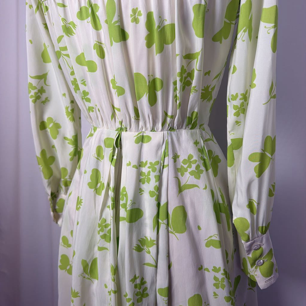 A white long-sleeve dress with a green botanical and butterfly pattern, featuring a gathered waist and tie detail, shown against a soft lavender background.