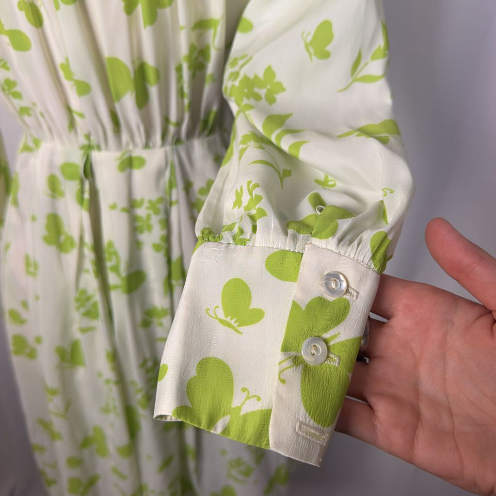 A close-up of a white long-sleeve blouse with a green butterfly and leaf print, featuring a buttoned cuff, shown with a hand holding the sleeve.