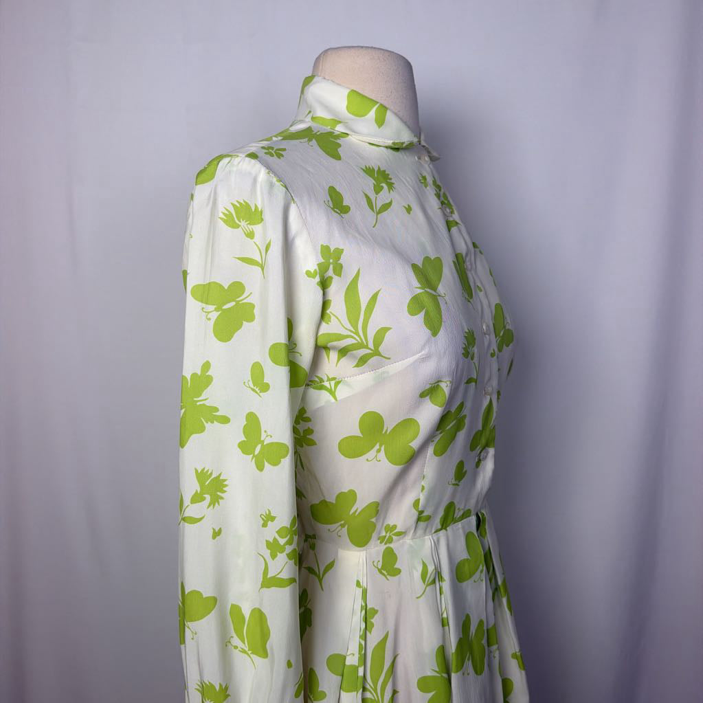 A long-sleeve collared dress displayed on a mannequin torso against a light gray fabric backdrop. The dress has an off-white base with a bright green floral and leaf print, a button front, and a self-tie waist belt.