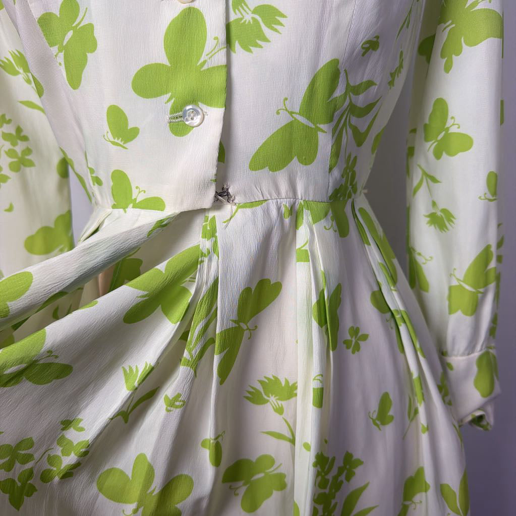 A long-sleeve collared dress displayed on a mannequin torso against a light gray fabric backdrop. The dress has an off-white base with a bright green floral and leaf print, a button front, and a self-tie waist belt.