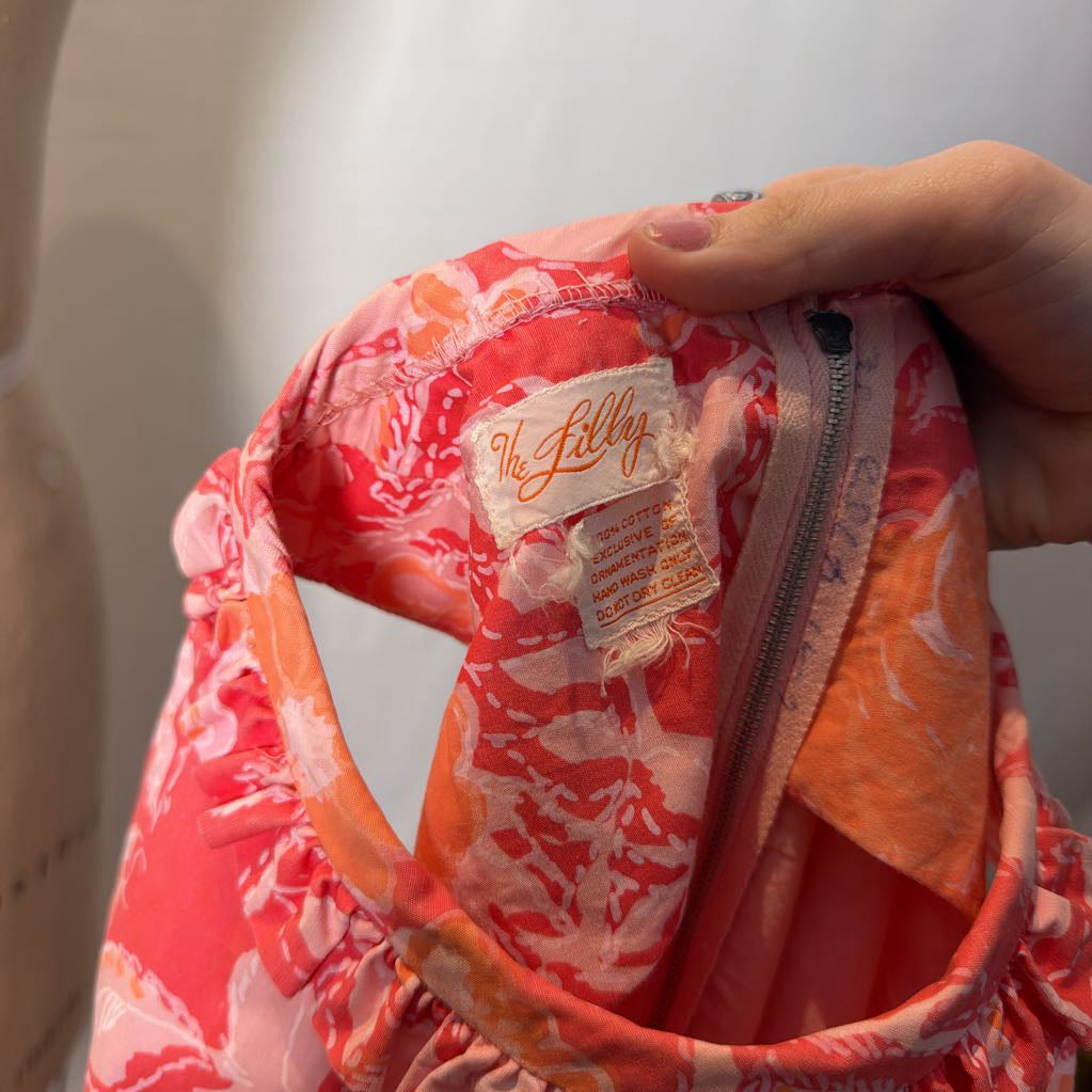 A hand holding a brand tag reading "the Lilly" on the inside of a dress in front of a white wall.