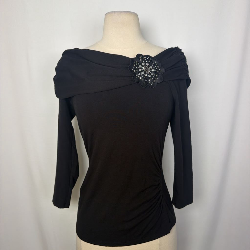 A black top with 3/4 sleeves and a draped collar with a large flower adornment on a mannequin in front of a white wall.