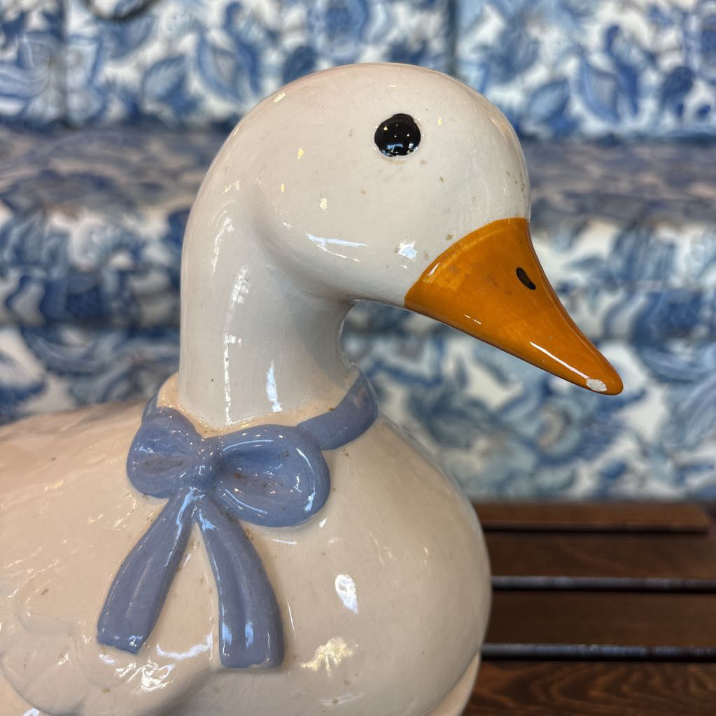 A ceramic white duck figure with a blue bow around its neck sitting on a wooden table in front of a blue and white sofa.