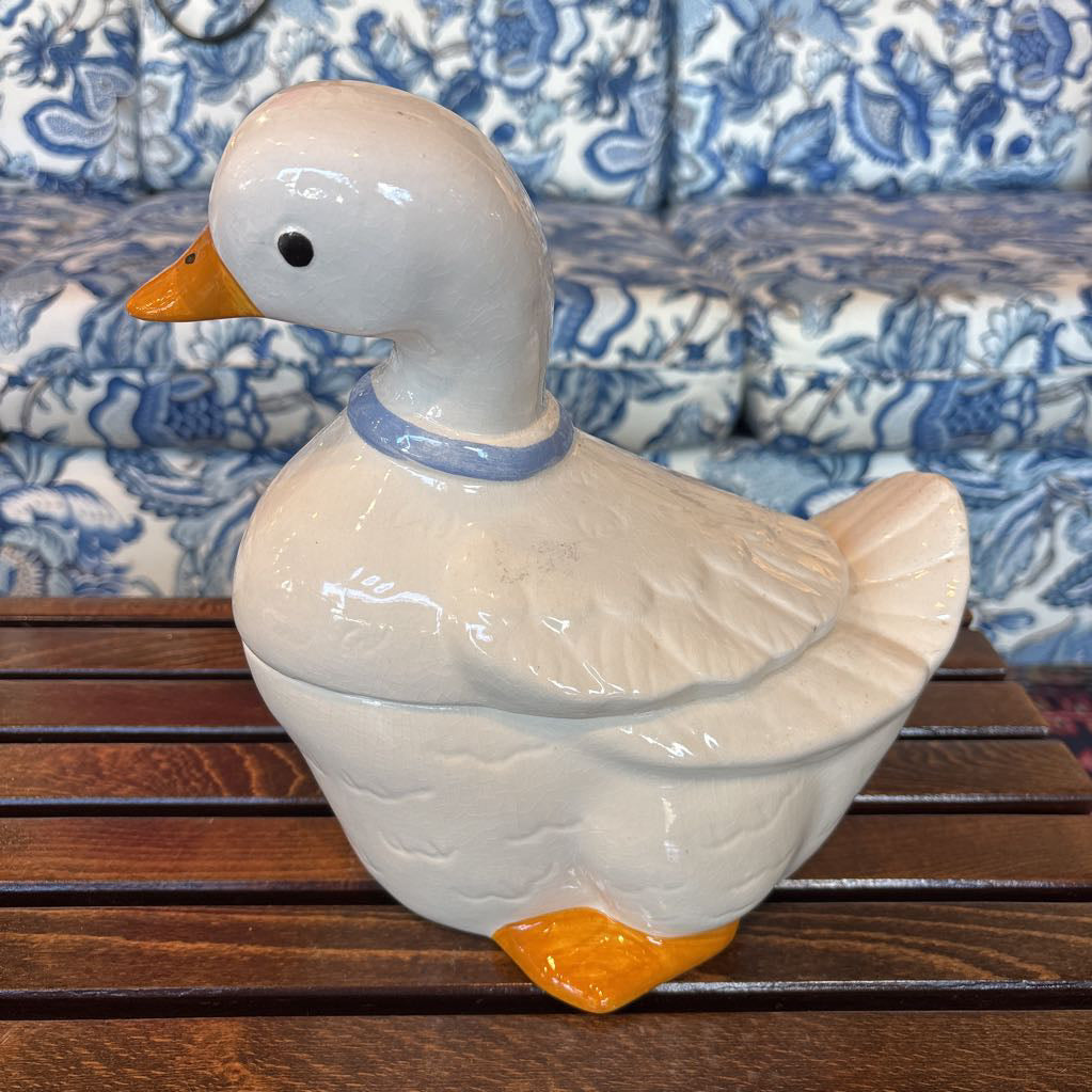 A ceramic white duck figure with a blue bow around its neck sitting on a wooden table in front of a blue and white sofa.