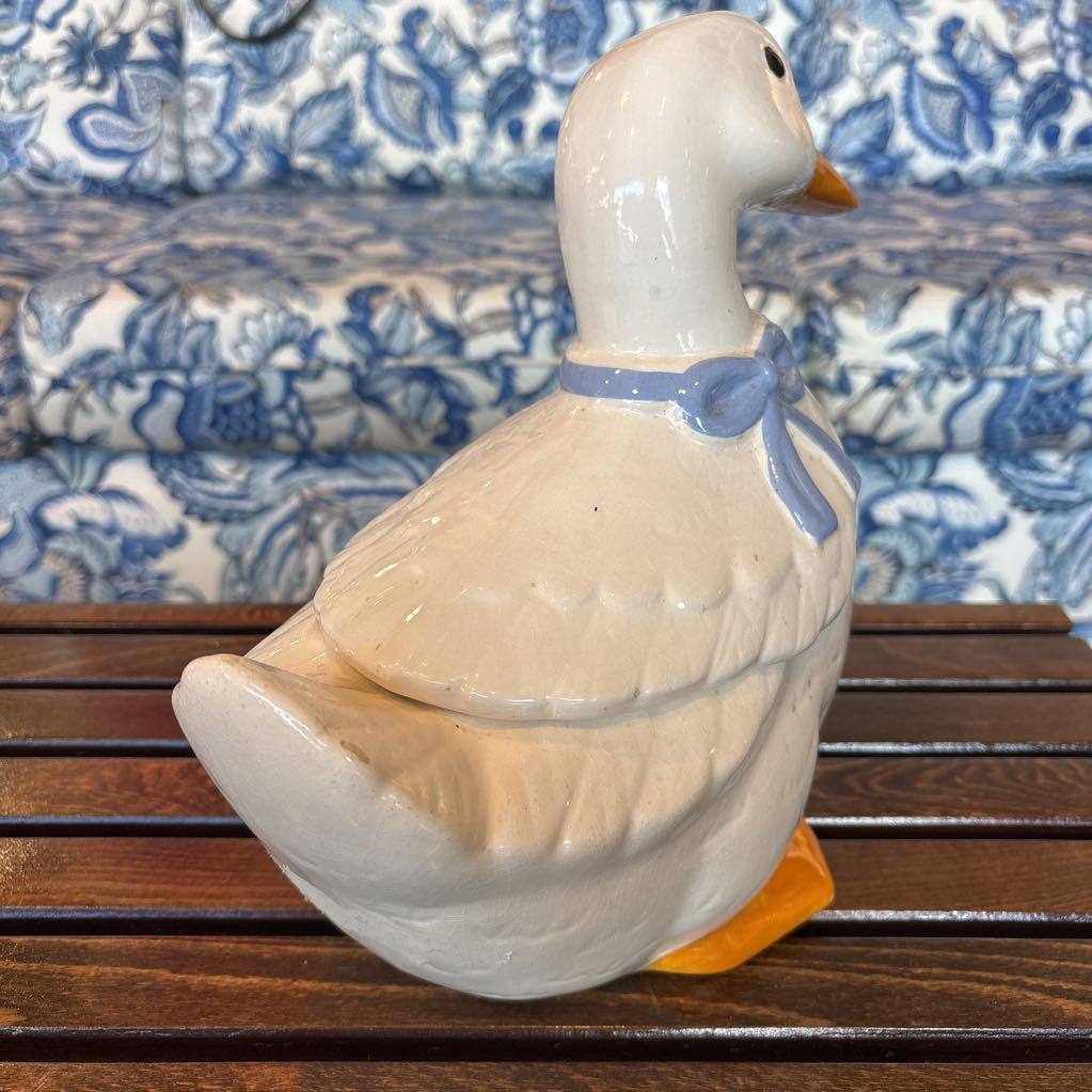 A ceramic white duck figure with a blue bow around its neck sitting on a wooden table in front of a blue and white sofa.