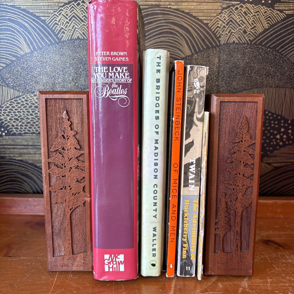 Two wooden bookends with laser cut images of tall trees on the front of them holding a stack of books. These sit on a wooden surface in front of a black and gold wallpaper.