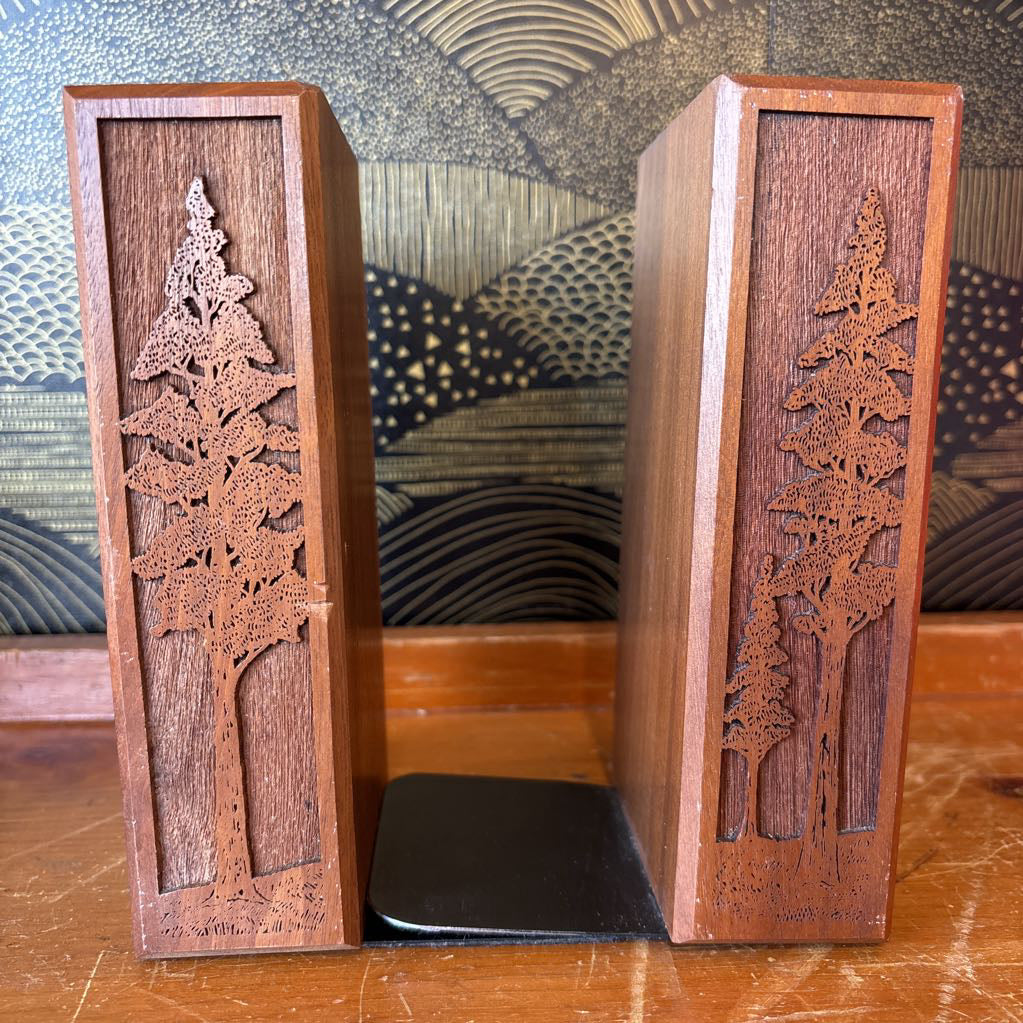 Two wooden bookends with laser cut images of tall trees on the front of them. These sit on a wooden surface in front of a black and gold wallpaper.