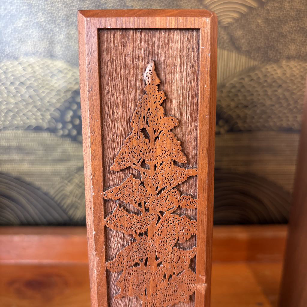 A wooden bookend with a laser cut image of a tall tree on the front sitting on a wooden surface in front of a black and gold wallpaper.
