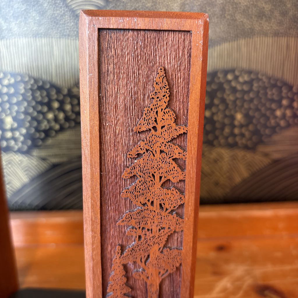 A wooden bookend with a laser cut image of a tall tree on the front sitting on a wooden surface in front of a black and gold wallpaper.